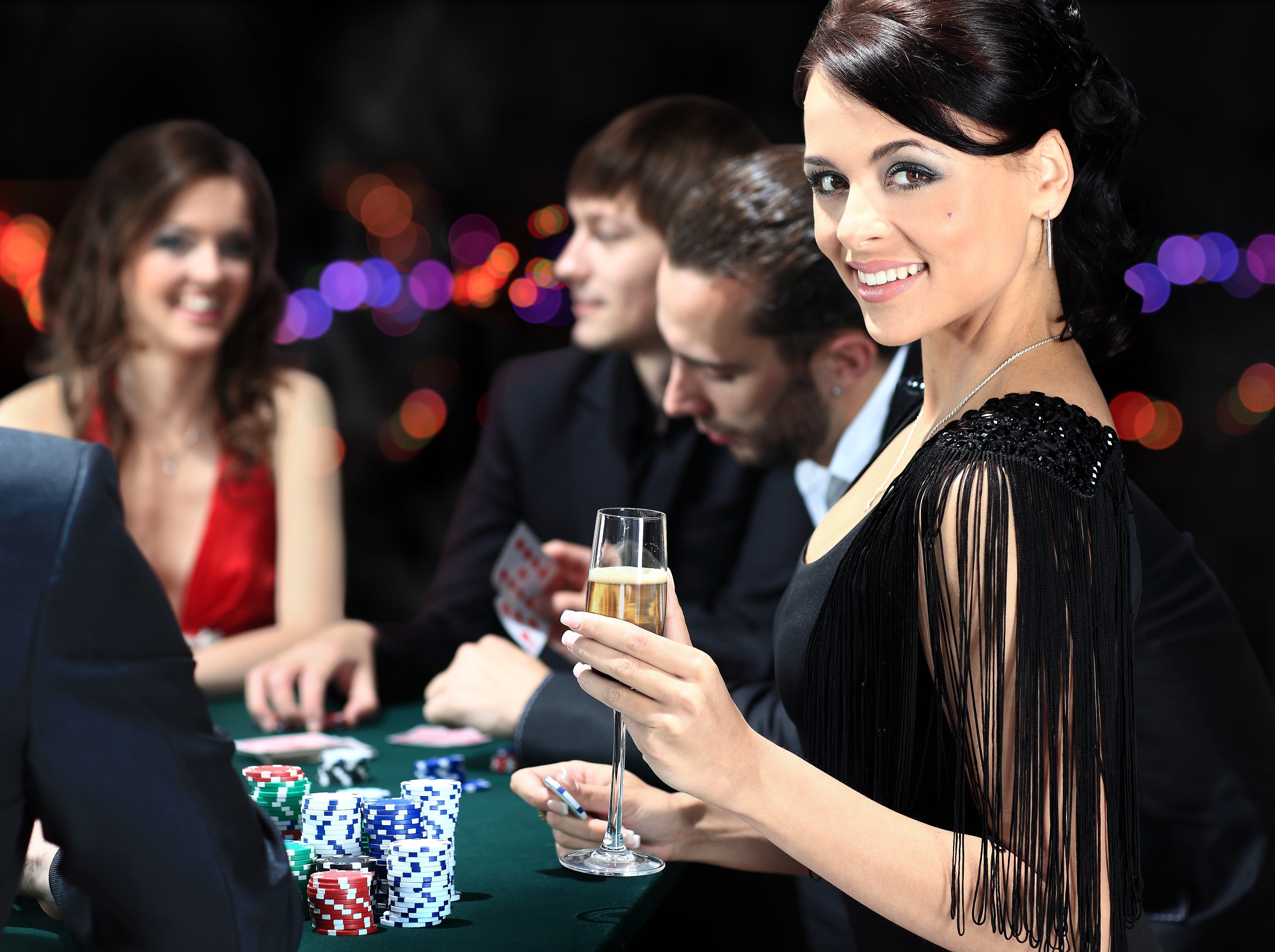 Брюнетка в оригинальном коктейльном платье позирует за игорным столом с бокалом в руках
