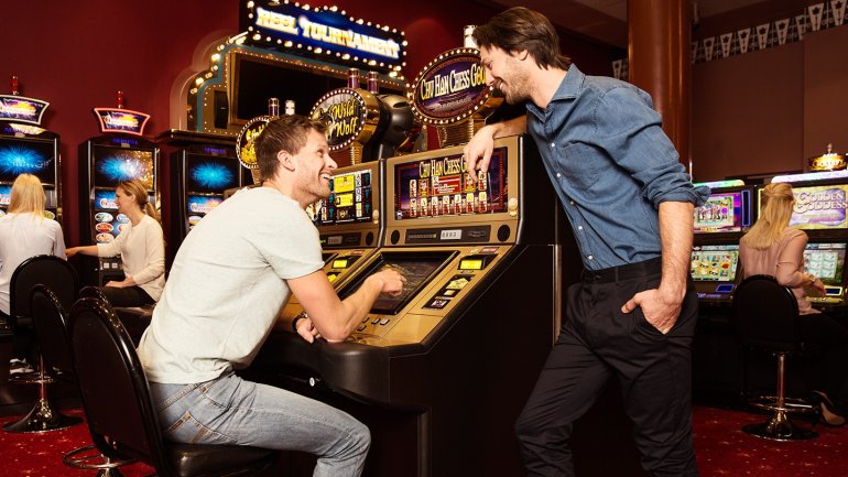 Молодой мужчина играет на автомате в зале казино, а его друг прислонился к аппарату и наблюдает за процессом