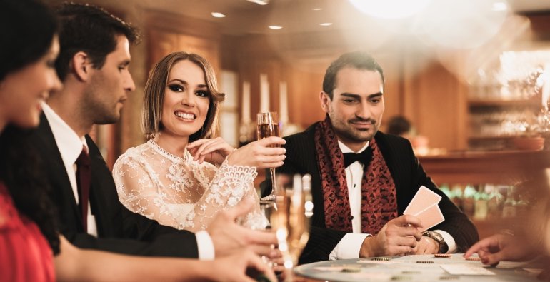 Элегантно одетые женщины и мужчины играют в казино, распивая шампанское