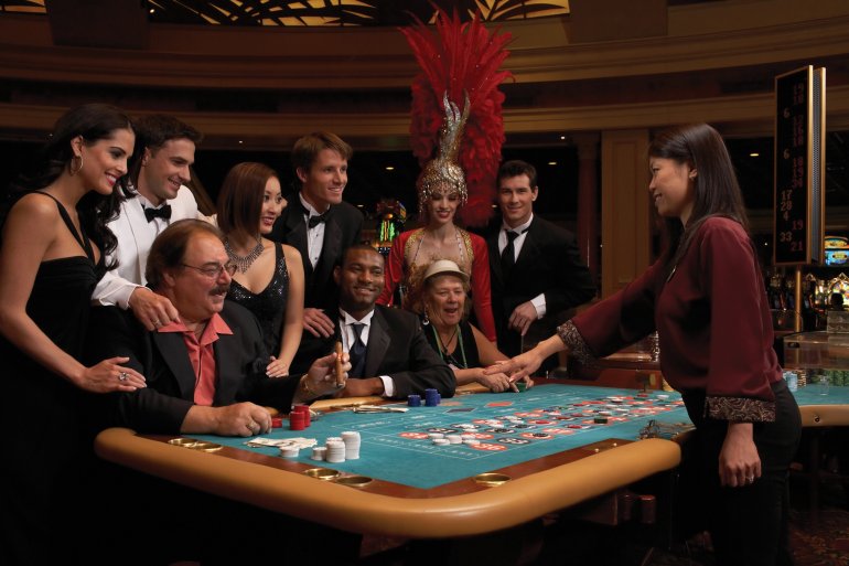 Элитные азартные игроки окружили стол для рулетки и развлекаются в процессе совершения ставок, а молодая темнокожая девушка крупье принимает ставки