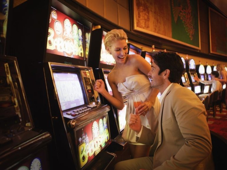 Женщина в вечернем платье и мужчина в строгом костюме играют в казино на игровом автомате за бокалом вина
