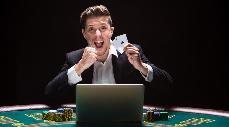 Молодой мужчина в строгом костюме сидит за покерным столом с картами в руках, а пред ним стоит ноутбук