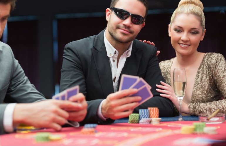 Мужчина в строгом костюме и затемненніх очках играет в покер, а рядом сидит его спутница, красивая блондинка в золотом вечернем платье