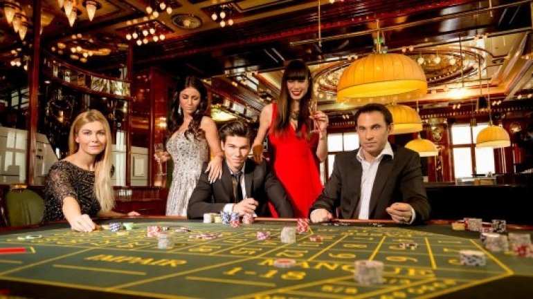 Трое женщин в роскошных вечерних платьях и мужчины в строгих костюмах веселятся и выпивают за игрой в казино VIP-уровня