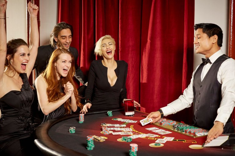 Молодой парень азиат ведет игру в блэкджек для веселой компании - троих женщин и молодого мужчины, которые во всю отрываются в казино