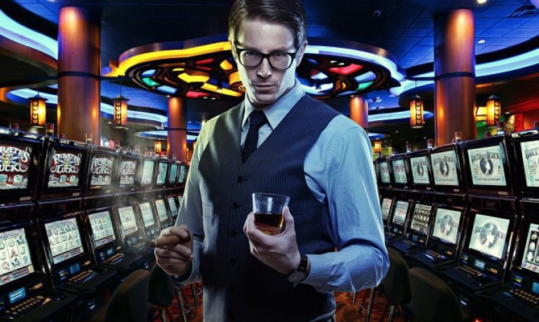 Загадочный молодой мужчина в очках и со стаканом виски стоит в центре зала игровых автоматов