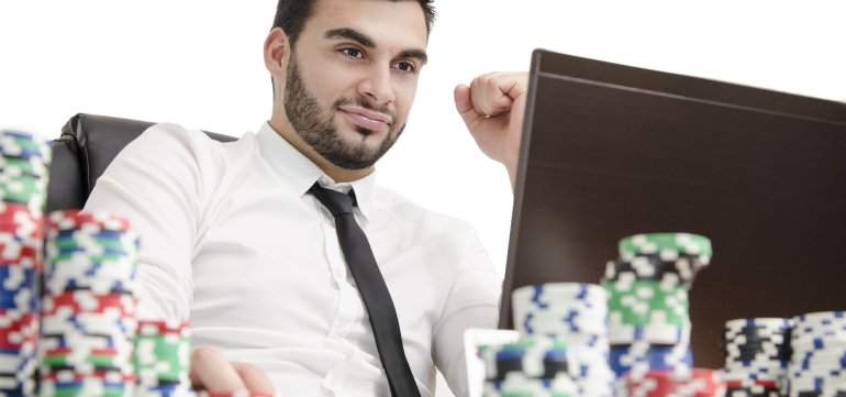 Молодой мужчина с легкой щетиной одетый в белую рубашку сидит за компьютером, а рядом стопки фишек для покера