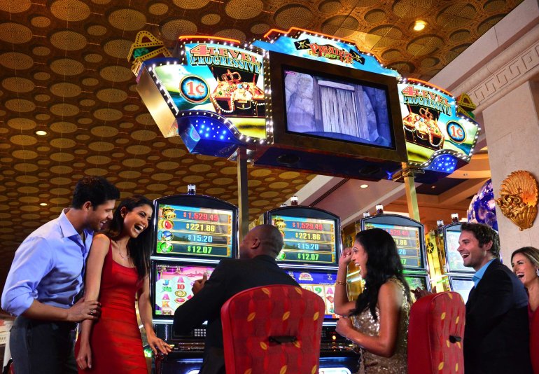 Две привлекательные брюнетки в красных вечерних платьях и трое мужчин в строгих костюмах веселятся за игровыми автоматами