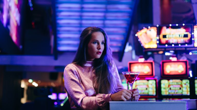Нарядно одетая молодая женщина скучает в зале с игровыми автоматами, распивая коктейль