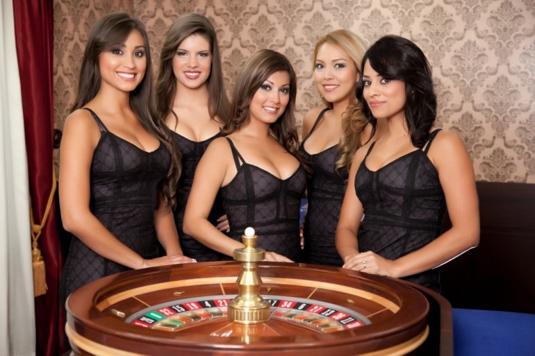 Пятеро сексуальных девушек крупье в обтягивающих черных платьях позируют возле стола для игры в рулетку